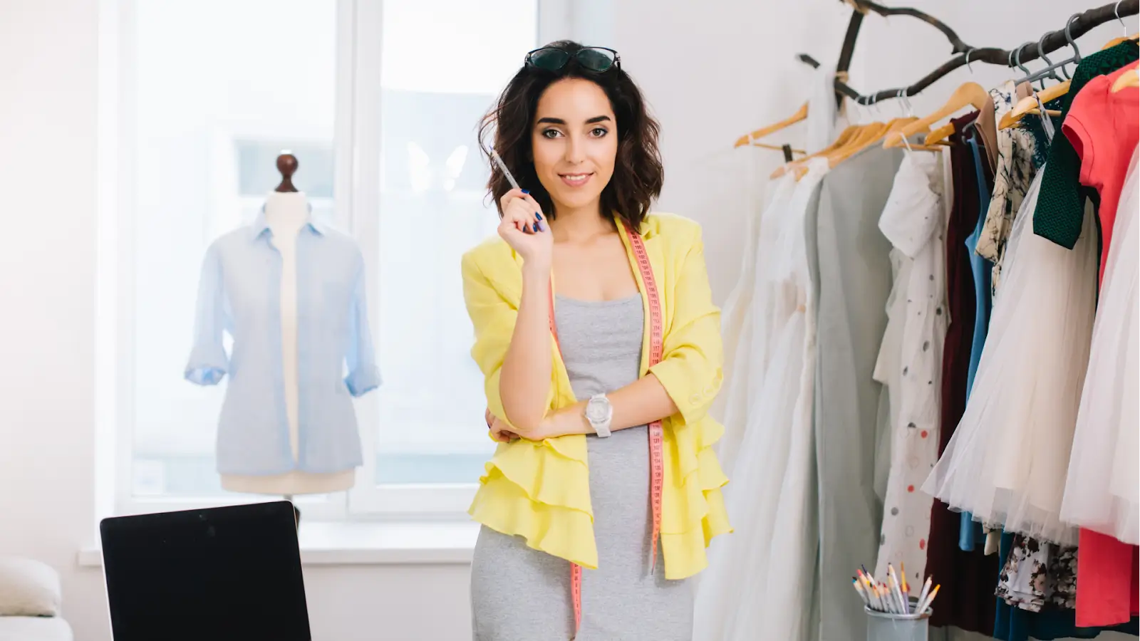 A stylist in a bright studio smiles at the camera, with a rack of clothes and a mannequin in the background.